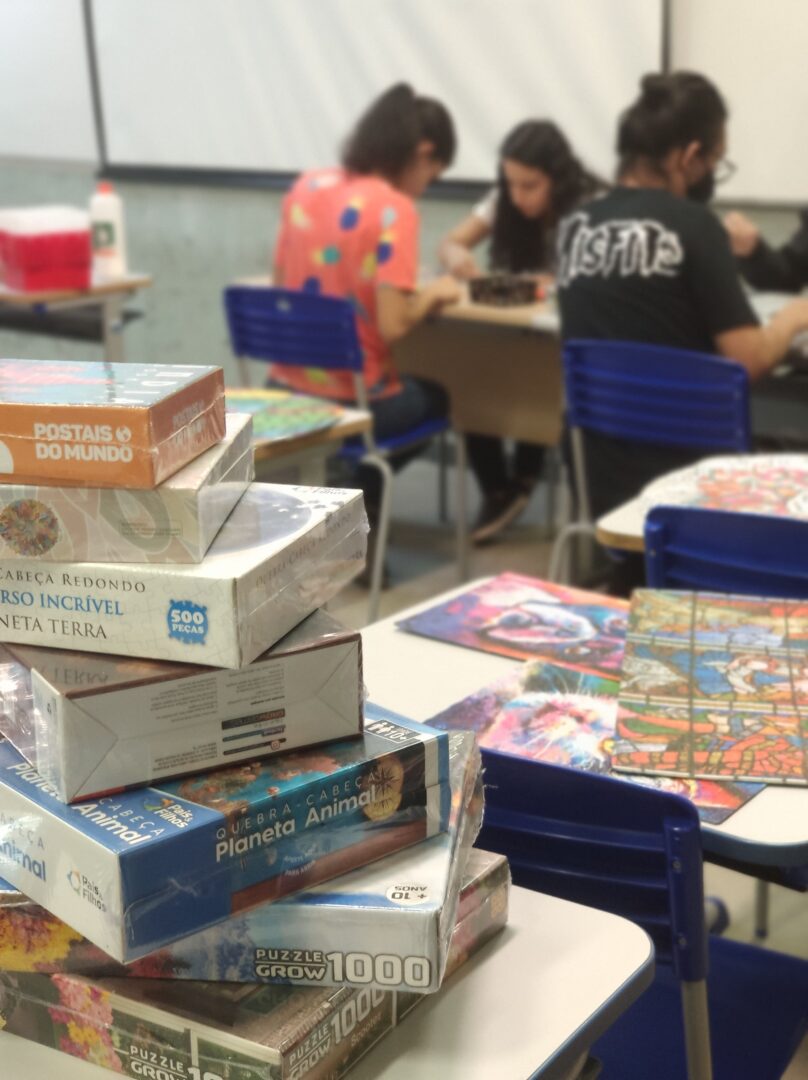 Uma sala com alunos, mesas espalhadas, cadeiras na cor azul e uma mesa com uma pilha de sete caixas de quebra-cabeça.