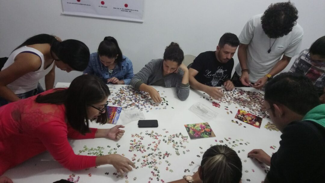 Uma sala com nove pessoas debruçadas em uma mesa tentando montar diversos quebra-cabeças coloridos juntas.